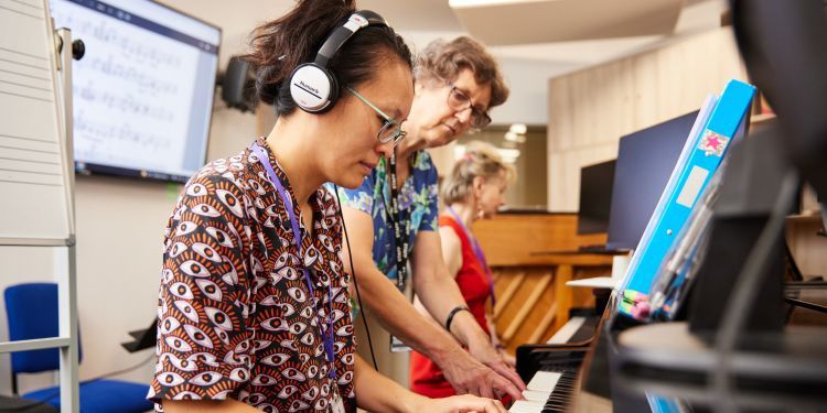 A woman sat at a keyboard wearing headphones. Aural teacher is sounds bass notes.