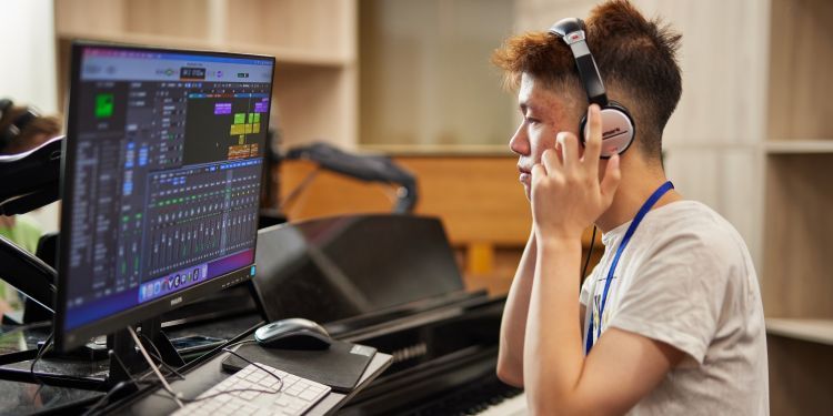 A man adjusting his headphones, sat at a laptop and piano keyboard working on music production software