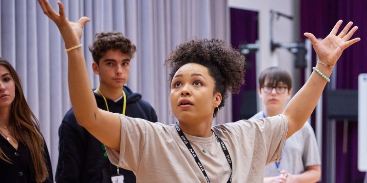 An acting teacher stood in a circle of students captures their attention, her arms raise aloft dramatically
