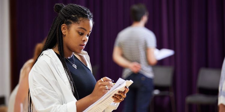 A woman with long braided hair standing in a rehearsal room, marking her script with a pencil