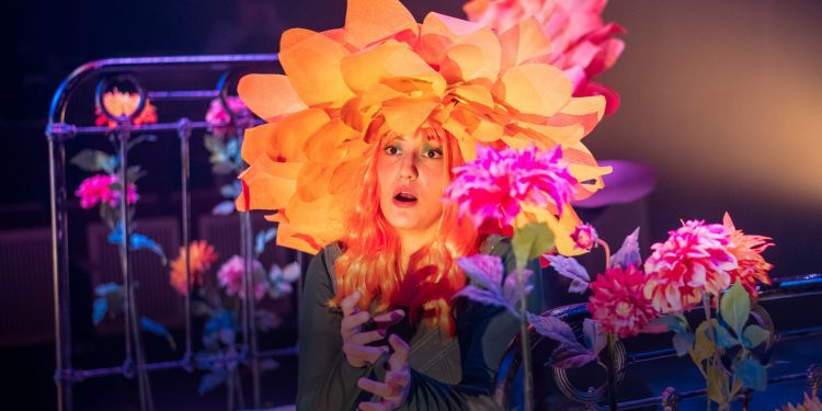 Guildhall opera singer dressed as flower, sitting on the edge of bed surrounded by colourful flowers