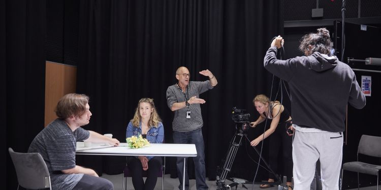 A male and female actor are sat at a table preparing to shoot a scene. The director is stood behind them, using his arms to instruct the camerawoman and boom operator how to frame the shot.