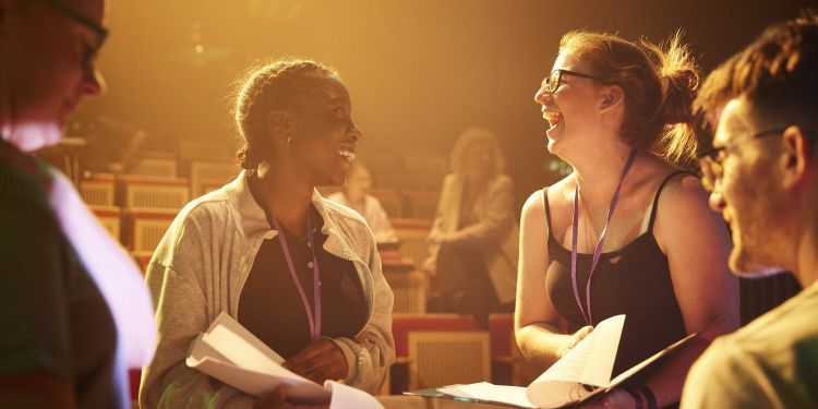 Two people standing up wearing lanyards in a theatre with clipboards and paper. They are smiling at each other.