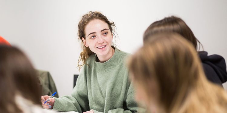 Picture of a woman sat down at a table with two people looking at them smiling, with a pen in her hand. Has has brown hair and is wearing a green jumper, and the background is white.