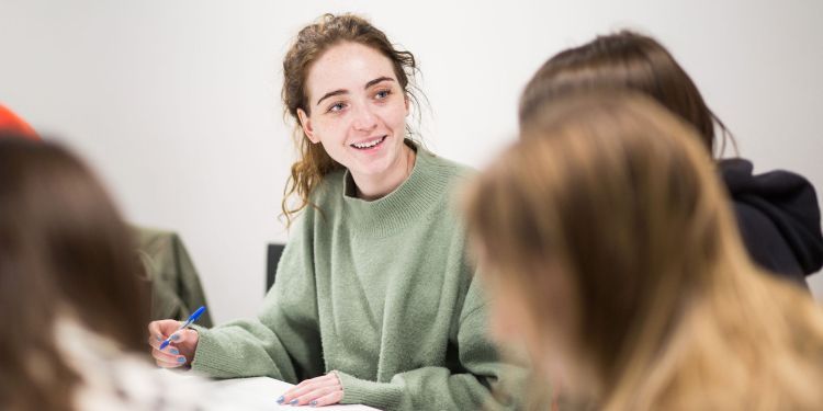 Picture of a woman sat down at a table with two people looking at them smiling, with a pen in her hand. Has has brown hair and is wearing a green jumper, and the background is white.