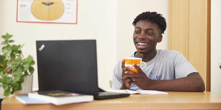 A boy holding a mug and sitting at a laptop 