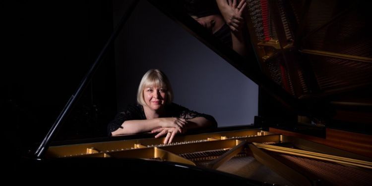 Woman with blonde hair sat behind piano with hands rested on the top of the piano