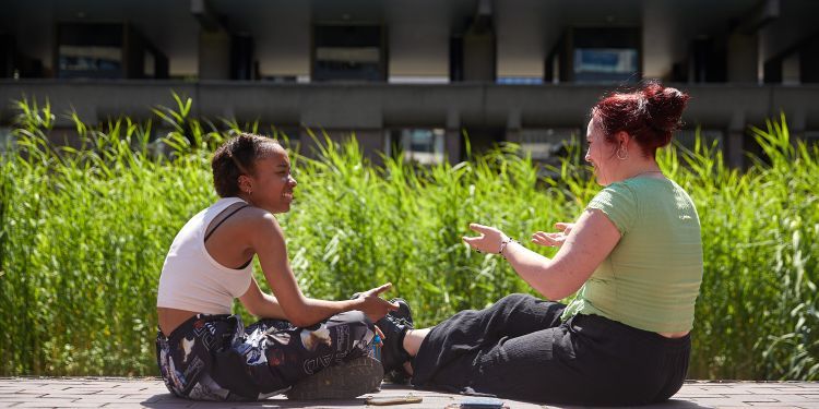 Two students laughing sat on the edge of the lake at Lakeside, Guildhall School