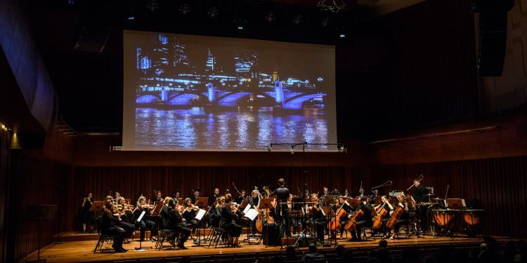 Orchestra on stage, with film showing in the background