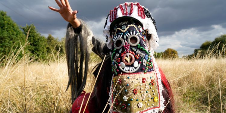 Person in decorative outfit and large mask standing in a field with long grass