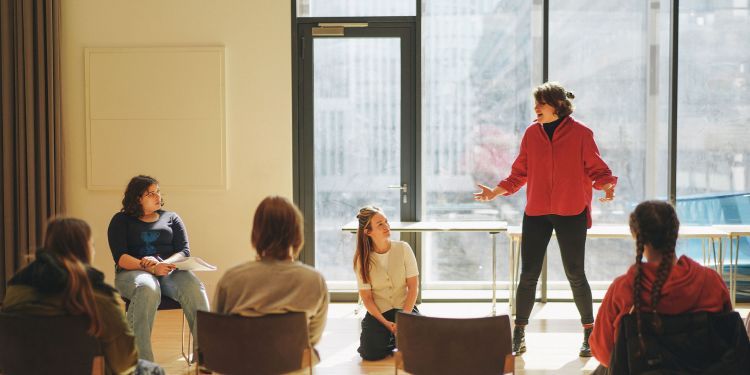 A woman in red stands next to two other women, one kneeling & one seated. She is directing a scene for the rest of the class who are sat on chairs facing her.