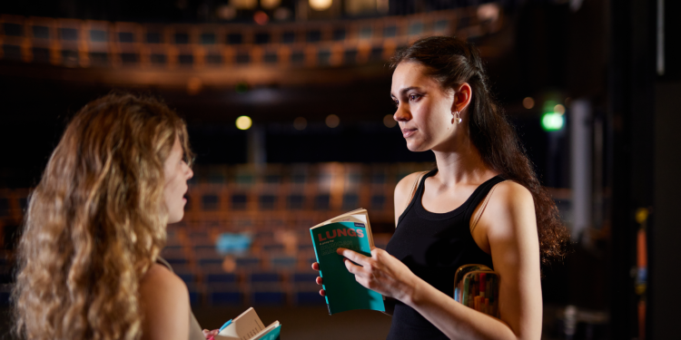 Image of two women in a theatre looking at each other with scripts in their hands