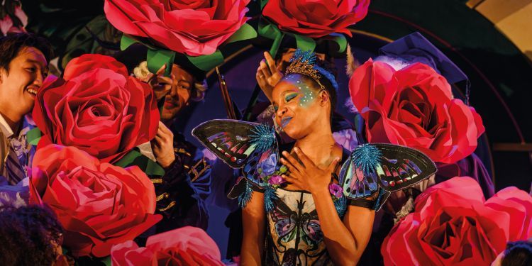 Performer wearing butterfly wings, tiara and colourful make up with hand on heart and eyes closed, surrounded by roses