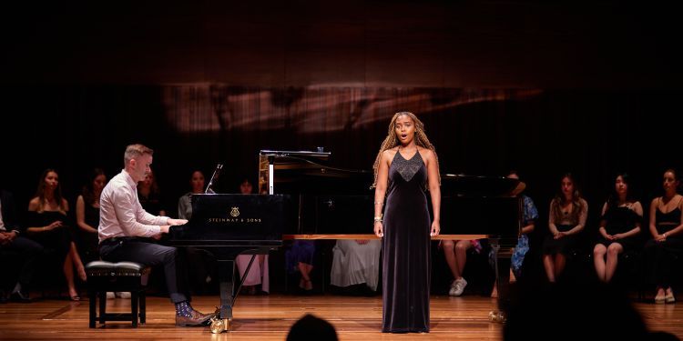 A young woman stands on stage, singing to an audience in a long black dress.