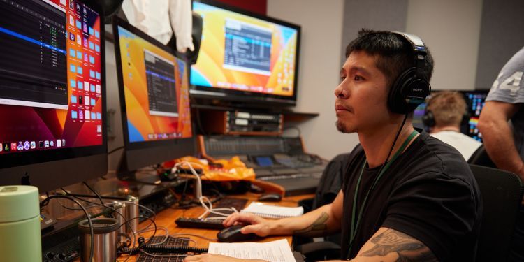 An Asian man sits in front of two computer screens at a desk, wearing a blank t-shirt and black headphones. On the screen he has two black windows open.