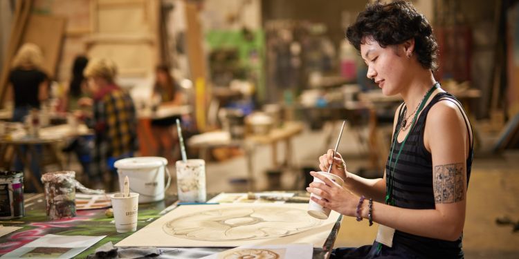 In the foreground towards the right, a person with short brown hair wearing a striped top and a tattoo on their arm sits at a desk with a paintbrush in one hand and a cup of water in the other. On the desk is a painting they are working on, as well as other paints and designs to copy from. In the background you can see other individuals at desks blurred in a studio.