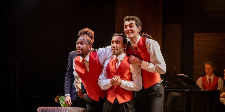 Three singers wearing red waistcoats and ties, and black trousers, with a grand piano in the background