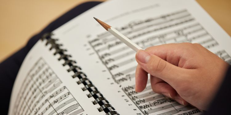 Close up image of a bounded book of sheet music for orchestra images. A white hand holding a white pencil rests on an open page.
