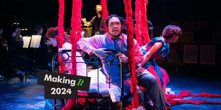 Three opera singers sat on a bed all facing different directions, with red ropes hanging down from ceiling. Small ensemble performing in background