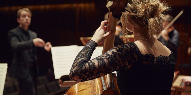 Student playing the double bass on stage with a conductor in the background