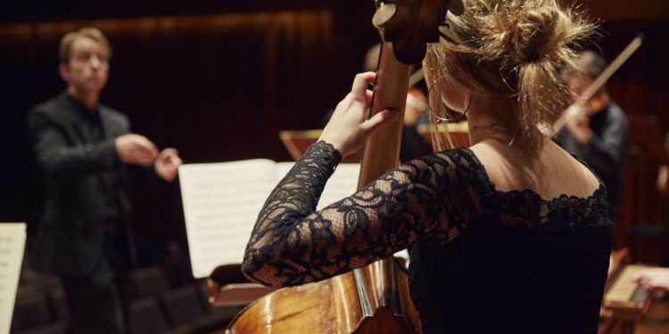 Student playing the double bass on stage with a conductor in the background 