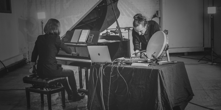 Two musicians where one is sat at a piano and the other is leaning over an electronic sound desk 