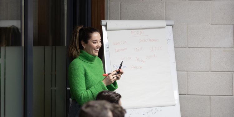 A tutor wearing a green jumper smiling in front of a flip chart