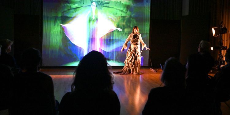 A woman sings in front of a screen showing her image, with an audience watching