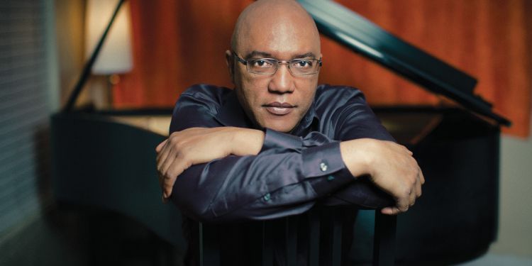 A man wearing glasses and a dark shirt leans on the back of a chair with a grand piano in the background.