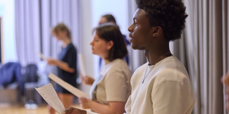 A row of people stand in a rehearsal room holding scripts, practising their speeches