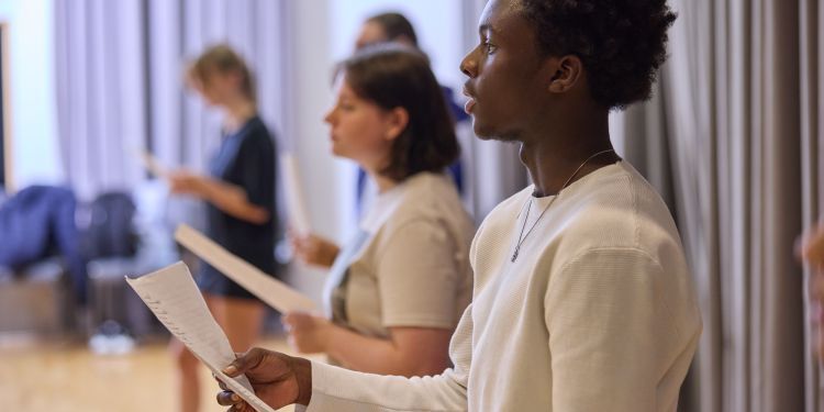 A row of people stand in a rehearsal room holding scripts, practising their speeches