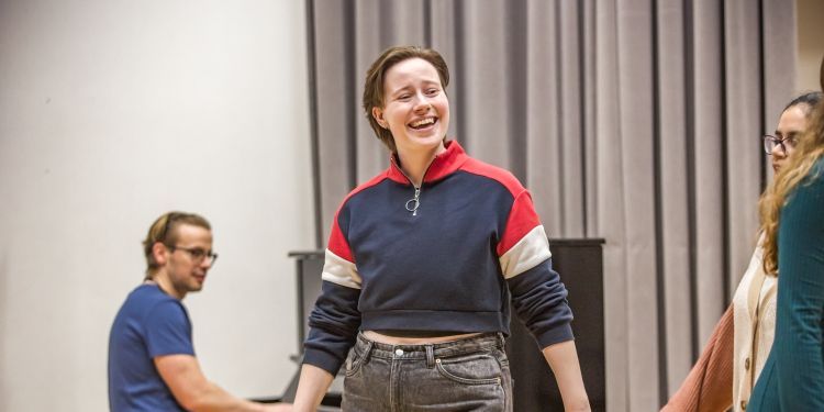 A smiling person moves through a rehearsal space with a pianist accompanying them in the background