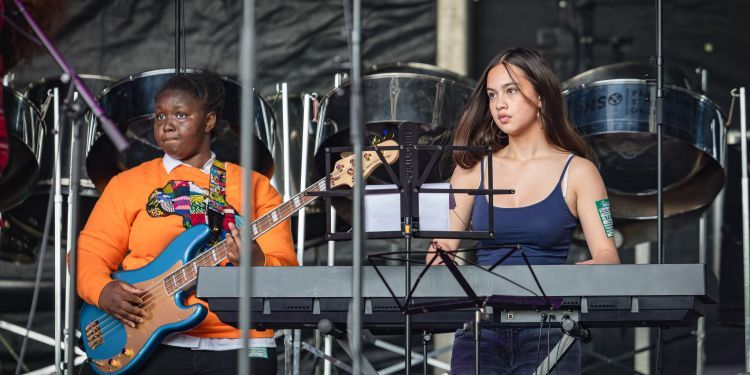 Two students perform on a stage. One wears an orange jumper and is playing a blue Precision bass guitar. The other wears a blue vest and plays the keyboard. There are steel pans in the background.