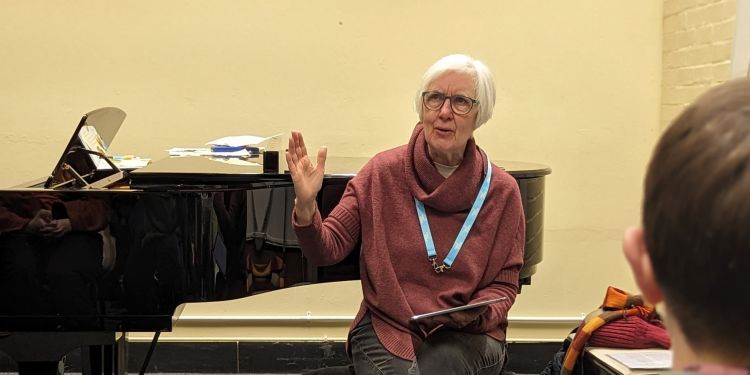 Dame Judith Weir sits in front of a black grand piano talking in a classroom. In the foreground there is the shoulder of a young boy in a grey jumper