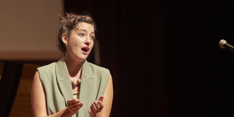 Vocal singer wearing a sage green short sleeved blazer singing on stage with their hands in front of them reaching out
