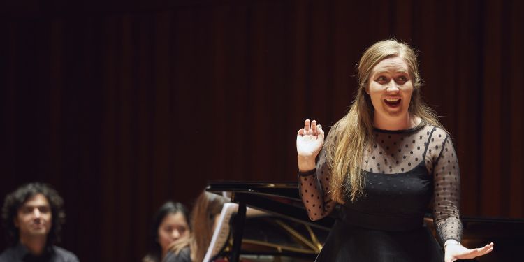 Vocal singer on stage mid performance wearing a black long sleeve dress, with two people in the background and a piano