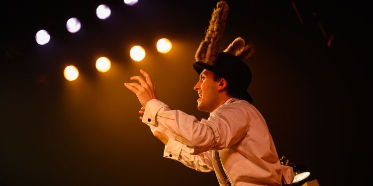 A student on stage wearing a white shirt and donkey fluffy ears on their head with their hands raised, facing upwards