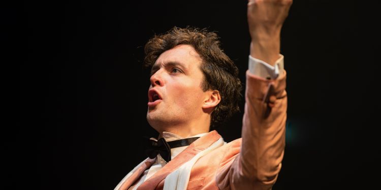 Opera student with shirt brown hair wear a pale pink suit and bow tie has one arm raised above their head whilst singing