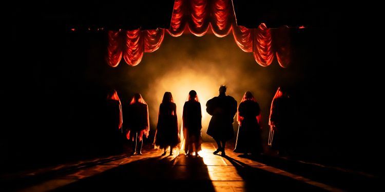 Production shot of Henry VIII and his 6 wives silhouetted in a warm yellow stage light with a red scalloped curtain framing the stage