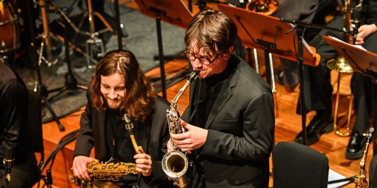 Student on stage playing the saxophone wearing a black tshirt and blazer with a student in the background sat down holding their saxophone