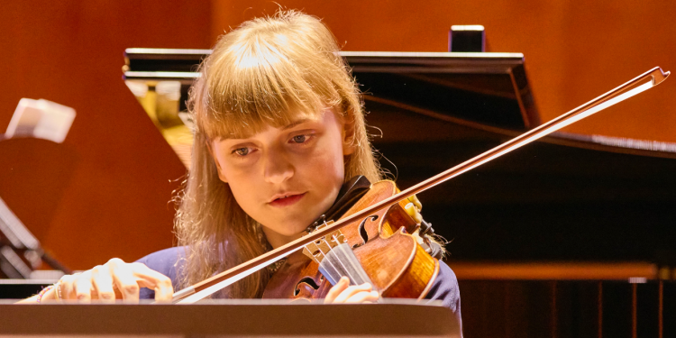 A girl with blond hair and a fringe plays the violin in a room with a dark wooden background and a grand piano behind her