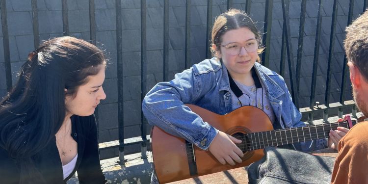 Two Music Makers students are sitting in the sun. The one on the right is holding a guitar. They are smiling while the workshop listener hears what they've been working on.