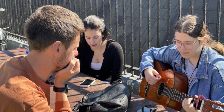 A tutor listens to two Music Makers students practicing a song they've been working on for the workshop. The three sit on a sunny rooftop terrace. 