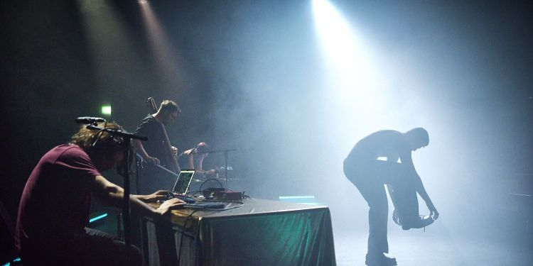 A dancer on stage performing with a spotlight on them with a student sat behind  a sound desk on a laptop to the left of the dancer