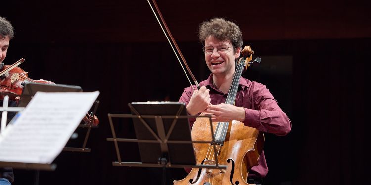Male presenting student laughing on stage holding their cello whilst seated