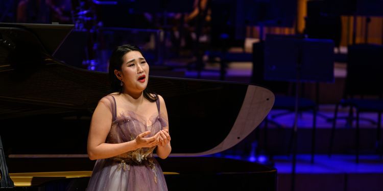 Female presenting singer on stage wearing a floor length purple evening gown singing in front of a piano