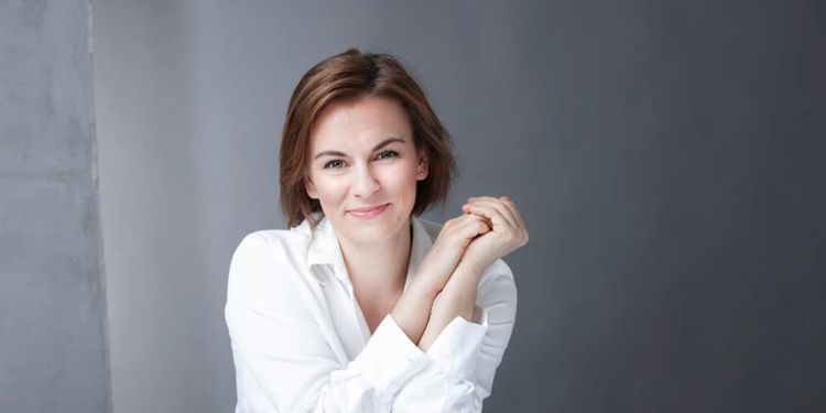 Conductor Daniela Candillari wearing a long sleeve white shirt sat on a white chair leaning against the back of it smiling with her hands together held near her face