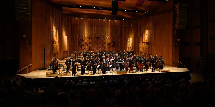 Guildhall Symphony orchestra on stage performing at Barbican Hall with an orange hue in the background 