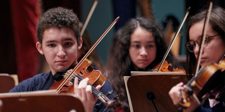 Three Junior Guildhall students on stage playing the violin concentrating and reading sheet music 