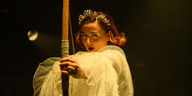 Opera student wearing a long tulle dress holding a bow and arrow aiming for the centre of the screen